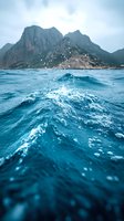 Dramatic ocean wave splashing against turquoise waters with rugged mountain peaks rising from misty coastline in background, creating moody seascape atmosphere.