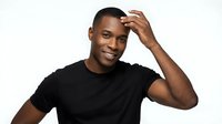 Young African American man with short dark hair wearing a black sleeveless shirt, smiling at camera with hand raised to head in casual, confident pose against white background.