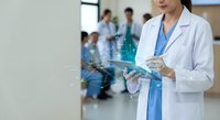Female healthcare professional in white lab coat using digital tablet in modern hospital corridor with medical team working in background.