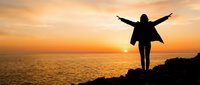 Woman silhouette with raised arms celebrating freedom on rocky coastline during golden sunset over ocean. Concept of success, achievement and life goals.