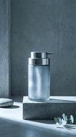 Modern glass soap dispenser with chrome pump on concrete surface in minimalist bathroom setting with natural lighting and eucalyptus leaves.