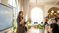 Professional businesswoman presenting financial data on whiteboard to diverse team in modern bright office with natural lighting and plants.