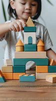 Young Asian girl playing with wooden building blocks in teal and orange colors, stacking and creating architectural structures on wooden table. Early childhood development.