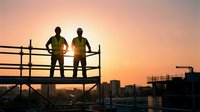 Two construction workers in safety vests standing on scaffolding platform at sunset overlooking urban development site with cranes and buildings.