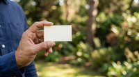 Man in blue shirt holding blank white business card outdoors with green foliage background for branding and corporate identity mockup.