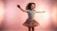 Young girl with curly hair jumping joyfully in striped shirt and pink skirt against soft pink background. Child expressing happiness and freedom in motion.