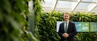 Professional businessman in dark suit standing confidently in modern greenhouse office with lush green plants and natural lighting for corporate sustainability.