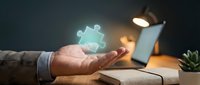 Business professional holding glowing puzzle piece above open book at desk with lamp, representing problem-solving and knowledge discovery in office.