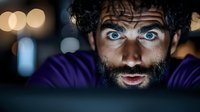Young bearded Caucasian man with intense blue eyes staring at computer screen in dark room, illuminated by display light, expressing shock and surprise at night.