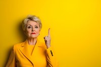 Confident mature blonde businesswoman in yellow suit pointing upward with finger against bright yellow background for corporate presentations.