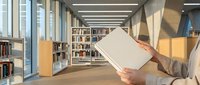 Person with light skin tone wearing a beige cardigan holds an open book in a modern library with wooden shelves, natural light, and contemporary architecture for educational and publishing content.