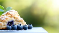Fresh baked scone with ripe blueberries on rustic surface against soft green bokeh background, close-up view showcasing morning pastry and fresh fruit.