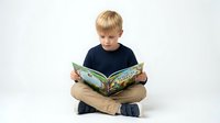 Young blonde Caucasian boy in navy shirt reading colorful storybook while sitting cross-legged on white background, education and childhood learning concept.