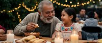 Elderly Caucasian grandfather and young Hispanic granddaughter sharing meal at outdoor family gathering with warm string lights creating cozy evening atmosphere.