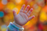 Small child's hand reaching up against blurred autumn bokeh background in warm orange and yellow tones, wearing cozy sweater sleeve. Represents innocence and wonder.