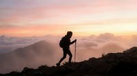 Silhouette of a hiker with trekking poles ascending a mountain ridge at sunrise above clouds, ideal for adventure and outdoor activity marketing.