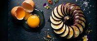 Fresh sliced eggplant arranged in circular pattern with cracked eggs on dark slate surface for healthy cooking and meal preparation concepts.