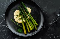 Fresh green asparagus spears with hollandaise sauce and herbs served on black plate against dark marble background, elegant restaurant presentation.