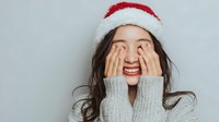 Young Asian woman in Santa hat covering eyes with hands while laughing joyfully, wearing cozy gray sweater against neutral background.