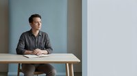Young professional man in casual shirt sitting at modern wooden desk in minimalist office space with soft natural lighting for business presentations.