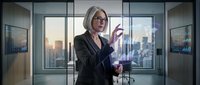 Professional blonde businesswoman in dark suit using holographic technology interface in modern glass office with city skyline view during sunset hours.
