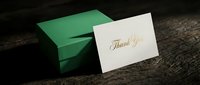 Elegant thank you card with gold foil script lettering leaning against a green gift box on a rustic dark wooden surface.