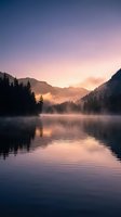 Serene mountain lake at dawn with misty fog and purple pink sky reflecting on calm water surrounded by forest hills for peaceful nature backgrounds.