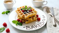 Stack of golden Belgian waffles topped with fresh raspberries, blueberries and mint on white plate with coffee cups.