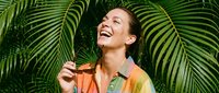 Happy Asian woman with dark hair laughing while holding fresh mango among tropical palm fronds, perfect for wellness, lifestyle, and summer content projects.