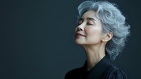 Serene mature Asian woman with silver gray hair and closed eyes against dark background, wearing black turtleneck, expressing peace and mindfulness.