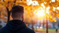 Young man in hoodie looking at golden autumn sunset through trees, back view silhouette against warm sunlight creating atmospheric outdoor scene.