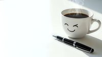 White coffee cup with cute smiling face next to black pen on clean desk surface with steam rising from hot beverage for morning productivity and workspace concepts.