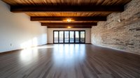 Empty spacious room with wooden floor, exposed brick wall, and rustic ceiling beams, illuminated by natural light through glass doors.