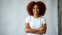 Confident young African American woman with curly hair smiling with arms crossed wearing white t-shirt against modern office background for business content.