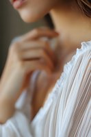 Close-up view of delicate white pleated fabric and feminine neckline, with soft focus on skin and gentle hand gesture, creating romantic and ethereal mood.