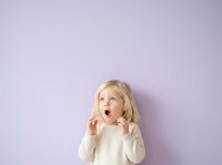 Young child with blonde hair and fair complexion, wearing a cream-colored long-sleeve shirt, displaying an excited or surprised expression with open mouth and raised hands against a soft lavender background.