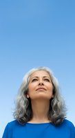 Mature woman with long gray hair wearing blue shirt looking upward against clear blue sky background.