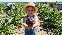Young girl in straw hat holding fresh purple eggplant at organic farm with workers harvesting vegetables in background during summer agricultural season.