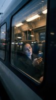 Woman sitting inside train window at night holding smartphone, looking out with contemplative expression. Evening commute, urban transportation, travel concept.