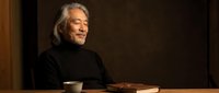 Elderly Caucasian man with gray hair and beard sitting at wooden table with coffee cup and books in warm indoor lighting for lifestyle content.