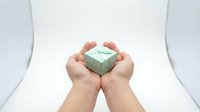 Hands gently holding small mint green gift box with white ribbon on clean white background. Concept of giving, protection, care and special occasions.