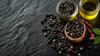 Black peppercorns scattered on dark slate with olive oil, dried chili peppers and fresh bay leaves, moody food photography with copy space.