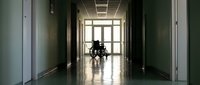 Person in wheelchair silhouetted in hospital corridor with natural light streaming through windows creating atmospheric healthcare environment.