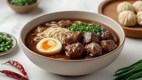 Asian beef noodle soup with tender meatballs, ramen, soft-boiled egg, and fresh herbs in ceramic bowl. Dark rich broth garnished with green onions and chili peppers.