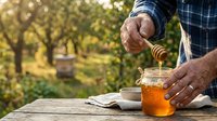 Middle-aged male beekeeper with tan skin pouring golden honey from wooden dipper into glass jar outdoors in orchard setting, wearing blue plaid shirt, representing apiary work and natural food production.