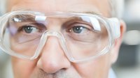 Close-up of senior Caucasian man wearing protective transparent safety goggles, focusing on blue eyes and upper face against blurred laboratory background.