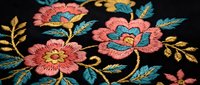 Colorful floral embroidery with pink and coral flowers on black fabric. Traditional needlework craft featuring decorative stitching and ornate botanical motifs.