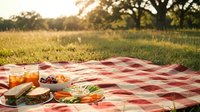 Outdoor picnic setup with food and drinks on red checkered blanket in sunny park meadow during golden hour sunset creating warm peaceful dining atmosphere.