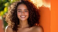 Young Latin American woman with natural curly hair and bright smile against orange wall background, radiating happiness and confidence in warm sunlight.