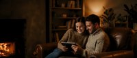 Young couple relaxing together on couch reading tablet in cozy living room with warm fireplace light creating intimate romantic atmosphere at home.